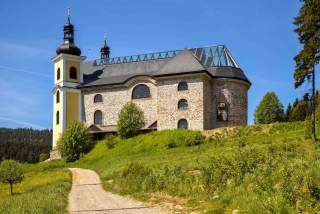 Eine Barockkirche mit teils verglastem Dach auf einer Bergwiese in der Sonne.