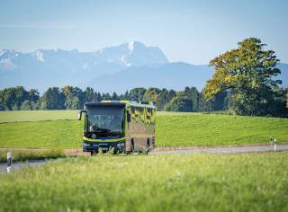 Ein moderner Bus fährt auf einer Landstraße durch grüne Felder, im Hintergrund sind Bäume und schneebedeckte Berge zu sehen.