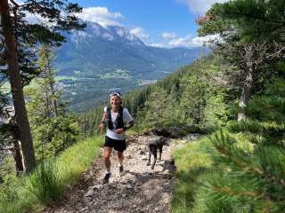Person in Sportkleidung läuft mit Hund auf einem felsigen Waldpfad, im Hintergrund Berge und blauer Himmel.
