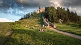 Eine Kirche auf einer Anhöhe, auf der rechten Seite Wald. Warmes Sonnenlicht bei gewittergrauen Wolken.