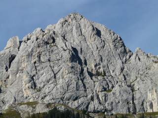 Großer, felsiger Berg mit einem Gipfelkreuz unter klarem blauem Himmel und einem bewaldeten Fuß.
