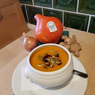 Weiße Schüssel mit orangener Kürbissuppe, garniert mit Kürbiskernen und Kürbiskernöl, daneben ein Kürbis, eine Zwiebel und Ingwer auf einem Holztisch.