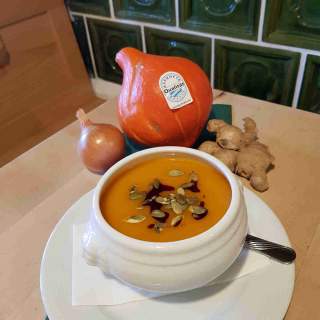 Weiße Schüssel mit orangefarbener Kürbissuppe, garniert mit Kürbiskernen und einem Faden Öl, daneben liegen eine Hokkaidokürbis, eine Zwiebel und ein Stück Ingwer.