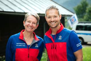 Frau Doktor Isabell Ruoß und Herr Henrik Vogel stehen vor ihrer Diensthütte. Sie tragen ihre typische blau-rote Kleidung. Im Hintergrund stehen Bäume und ein Rettungsfahrzeug.