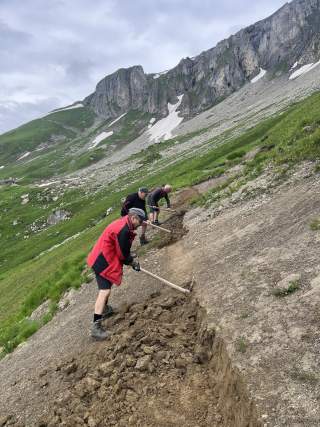 Drei Männer stehen an einem Berghang und hacken die Erde, um einen Bergweg in den Hang zu schlagen.
