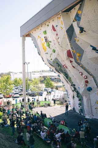 Außenaufnahme einer Kletterwand mit bunten Griffen, an der Sportler klettern, während unten zahlreiche Zuschauer auf Bänken sitzen und das Geschehen verfolgen.