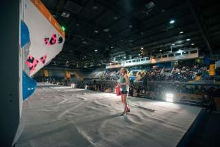 Eine Kletterin steht auf einer Boulder-Wettkampffläche vor einer Kletterwand mit bunten Griffen, im Hintergrund sitzt ein großes Publikum in einer Sporthalle.