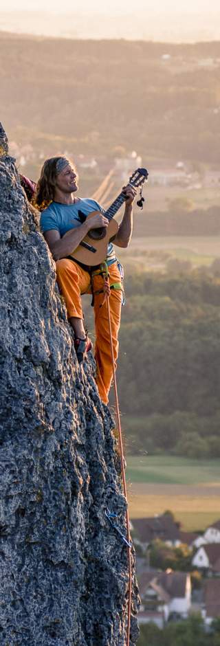 Ein Kletterer sitzt gesichert an einer Felswand und spielt Gitarre, im Hintergrund ist eine Landschaft mit Feldern und Häusern zu sehen.