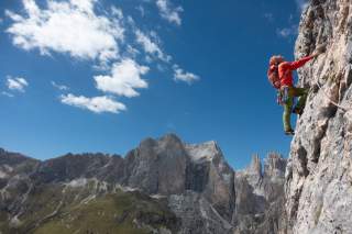 Ein Kletterer mit Helm und Ausrüstung klettert eine steile Felswand, im Hintergrund Berge unter blauem Himmel mit wenigen Wolken.
