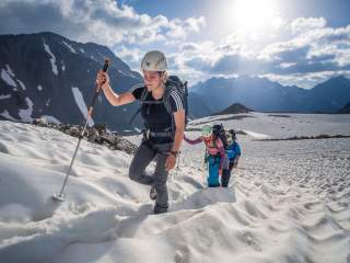 Drei Bergsteiger mit Helmen und Rucksäcken steigen bei sonnigem Wetter eine schneebedeckte Gletscherfläche in den Bergen hinauf.