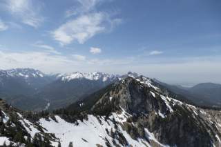Berglandschaft mit schneebedeckten Gipfeln und grünen Wäldern unter einem blauen Himmel mit vereinzelten Wolken.