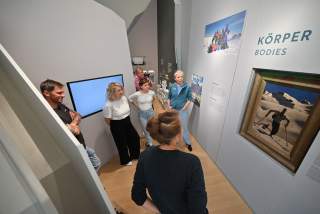 Eine Gruppe von Menschen steht in einem Ausstellungsraum vor einer Wand mit Bildern und Texten zum Thema „Körper“ und betrachtet die Exponate.