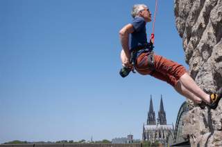 Ein älterer Mann klettert an einer künstlichen Kletterwand, im Hintergrund sind der Kölner Dom und die Hohenzollernbrücke zu sehen.