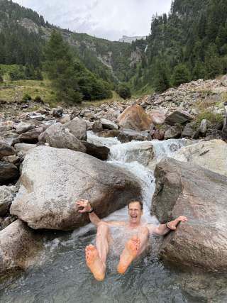 Ein lachender Mann sitzt in einem Gebirgsbach zwischen großen Felsen, umgeben von grüner Berglandschaft.