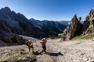Eine Wanderin und und ein Wanderer mit großen Rucksäcken steigen ein Schuttkar zwischen erodierten Kalkgipfeln empor.