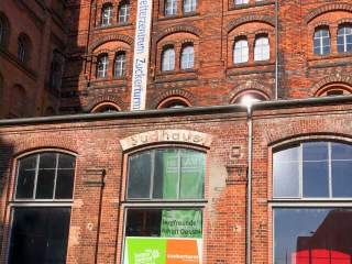 Backsteinfassade eines alten Industriegebäudes mit großen Fenstern; an der Wand steht „Kletterzentrum Zuckerturm“, Banner und Schilder weisen auf eine Kletterhalle hin.