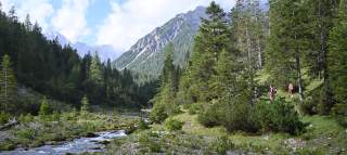 Berglandschaft mit Nadelwald, einem Bach im Vordergrund und Wanderern auf einem Pfad. Im Hintergrund ragen Berge unter leicht bewölktem Himmel auf.