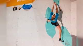 Ein Boulderer hängt an einem blauen Griff an einer Kletterwand bei der Deutschen Meisterschaft Bouldern 2025. Die Wand ist mit weiteren bunten Griffen in Gelb und Beschriftungen versehen.