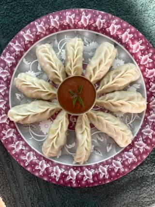 Ein Teller mit zwölf gedämpften Momos, die kreisförmig um eine Schale mit roter Sauce angerichtet sind.