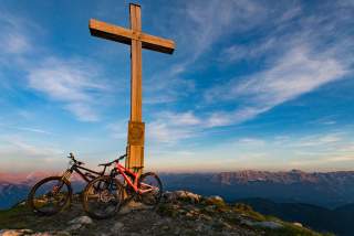 Zwei Mountainbikes lehnen an einem großen Holzkreuz auf einem Gipfel mit Blick auf ein Bergpanorama und einen blauen Himmel bei Sonnenaufgang oder Sonnenuntergang.