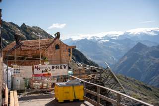 Bauarbeiter decken das Dach eines Holzhauses auf einer Baustelle in den Bergen, umgeben von einem Geländer und mit schneebedeckten Gipfeln im Hintergrund.