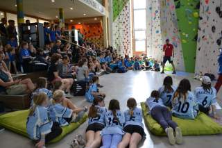 Viele Kinder und Jugendliche sitzen in einer Kletterhalle auf Matten und Stufen und schauen zu einem Mann mit Mikrofon, der vor einer Kletterwand steht.