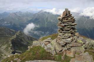 Gestapelte Steine auf einem Berggipfel mit Blick auf bewaldete Berge und Wolken im Hintergrund.
