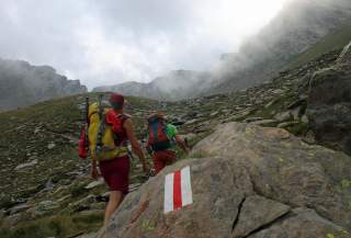Zwei Wanderer mit großen Rucksäcken steigen auf einem felsigen Gebirgspfad bei nebligem Wetter auf, vor einem Stein mit einer rot-weiß-roten Wegmarkierung.