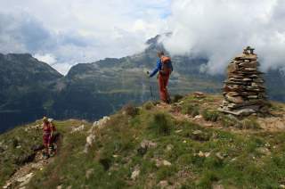 Zwei Wanderer mit Rucksäcken und Trekkingstöcken auf einem Bergpfad mit Gipfelkreuz aus Steinen, umgeben von bewölkten Bergen.