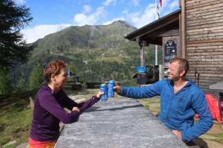 Zwei Personen stoßen mit Dosen an einem Steintisch vor einer Berghütte in einer alpinen Landschaft an.