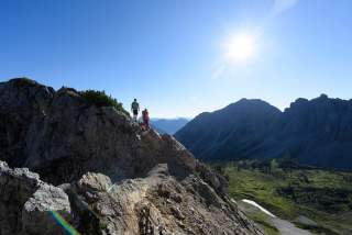 Zwei Wanderer stehen auf einem felsigen Berggrat mit Blick auf ein Tal und dahinter auf hohe, schroffe Berggipfel bei sonnigem, klarem Himmel.