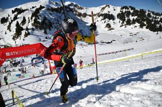 Winterlicher Skibergsteiger in bunter Sportkleidung und Helm erklimmt mit Skistöcken einen verschneiten Hang vor einer Bergkulisse mit blauem Himmel.