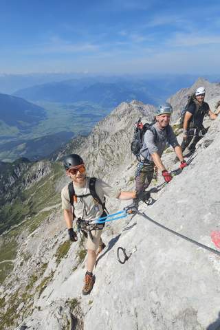 Drei Männer klettern gesichert an einer steilen Felswand in den Bergen bei klarem Himmel, im Hintergrund sind grüne Täler und Bergketten sichtbar.
