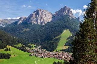 Ein kleines Dorf liegt eingebettet in grünen Wiesen und Wäldern am Fuße imposanter, felsiger Berge unter blauem Himmel.
