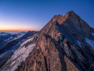 Bergsteiger mit Helm und Ausrüstung klettert bei Sonnenaufgang auf einem felsigen Berggrat, im Hintergrund schneebedeckte Gipfel.