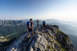 Zwei Wanderer mit Rucksäcken gehen auf einem felsigen Berggipfel mit einem Kreuz, umgeben von einer weiten Berglandschaft unter klarem Himmel.