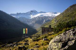 Eine Steinhütte schmiegt sich an einen Grashang davor zwei bunte Fahnen und im Hintergrund ein großer Gletscher