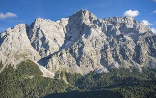 Felsige Bergspitzen mit steilen Klippen unter blauem Himmel, darunter ein bewaldeter Hang.