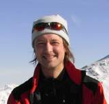 Porträt eines lächelnden Mannes mit Mütze und Sonnenbrille auf dem Kopf, der in einem roten und schwarzen Wintermantel vor schneebedeckten Bergen steht.