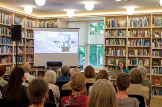 Vortragsveranstaltung in einer Bibliothek mit sitzendem Publikum, einer Frau am Rednerpult und einer Präsentation auf einer Leinwand.