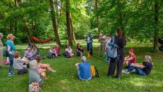 Gruppe von Erwachsenen sitzt und steht im Kreis auf einer Wiese im Wald, während eine Person in der Mitte aus einem Buch vorliest.