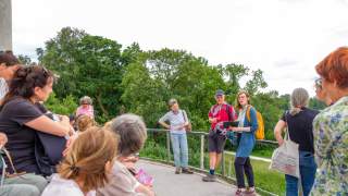 Gruppe von Menschen steht und sitzt im Freien vor grünem Baumbestand, eine Frau mit gelbem Rucksack spricht zur Gruppe.