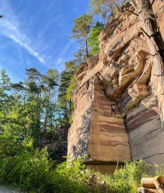 Person klettert an einer hohen Felswand in einem bewaldeten Gebiet unter blauem Himmel.