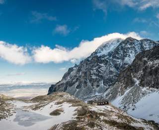 Schneebedeckte Berglandschaft mit steilen Felsen und einer Berghütte am Hang unter blauem Himmel mit wenigen Wolken.