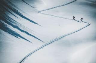 Zwei Skitourengeher gehen eine verschneite Hangfläche hinauf und hinterlassen eine geschwungene Spur im Schnee.
