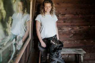 Eine Frau mit lockigen Haaren steht in einem Holzraum, lächelt und schaut auf einen schwarzen Hund, der sie ansieht; im Bild ist auch ihr Spiegelbild zu sehen.