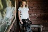Eine Frau mit lockigen Haaren steht in einem Holzraum, lächelt und schaut auf einen schwarzen Hund, der sie ansieht; im Bild ist auch ihr Spiegelbild zu sehen.