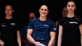 Fünf junge Frauen stehen vor schwarzem Hintergrund, die im Vordergrund stehende Frau trägt ein blaues Marmot-Shirt und hält eine Chalkbag, während sie lächelt. Die anderen Frauen tragen Sportkleidung und blicken in verschiedene Richtungen.