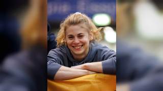 Junge Frau mit blondem Haar und grauem Hoodie lehnt lächelnd mit verschränkten Armen auf einem gelben Gegenstand.