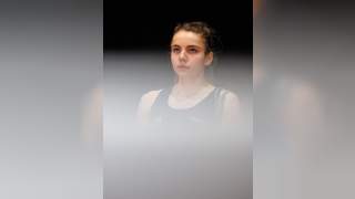 Porträt einer jungen Frau mit hochgebundenem Haar und schwarzem Sporttop vor dunklem Hintergrund, teilweise von unscharfem, weißem Vordergrund bedeckt.